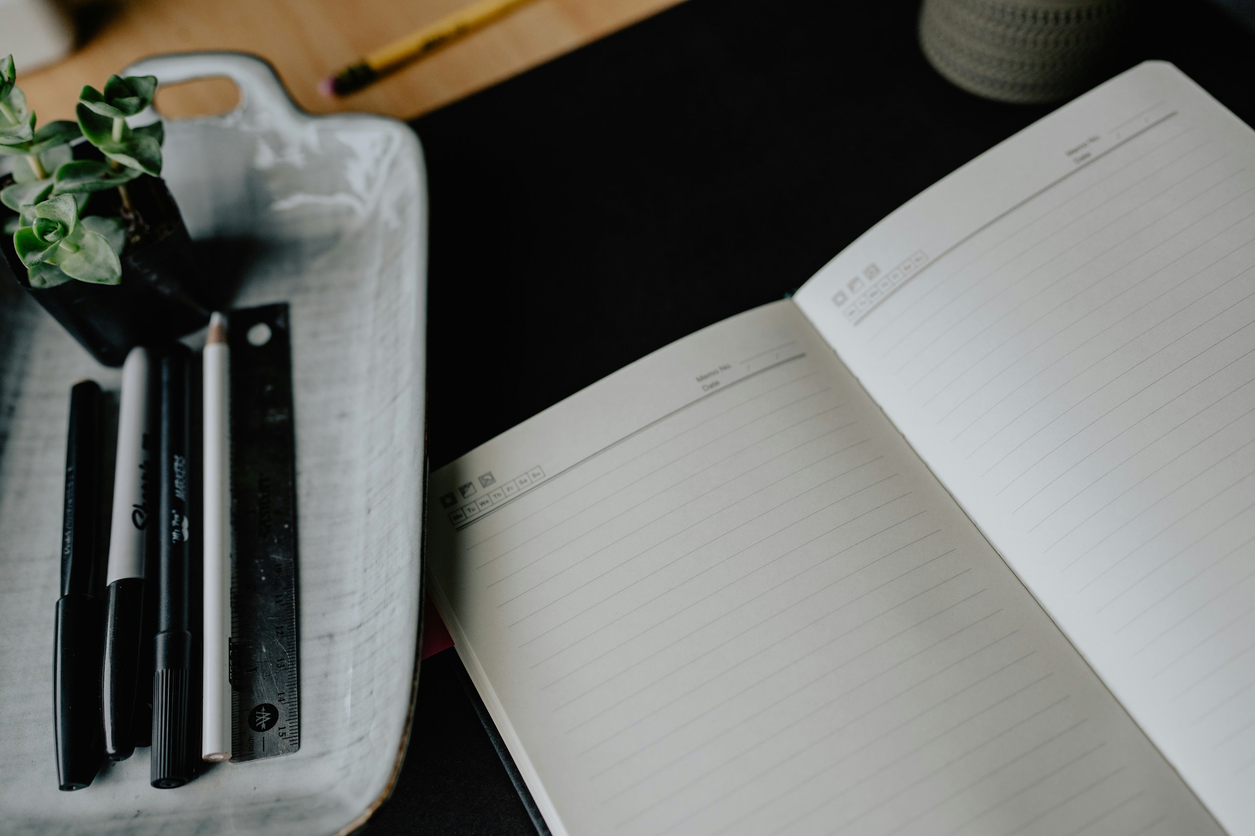 An open, blank lined notebook on a dark desk beside a tray of pens and a small succulent