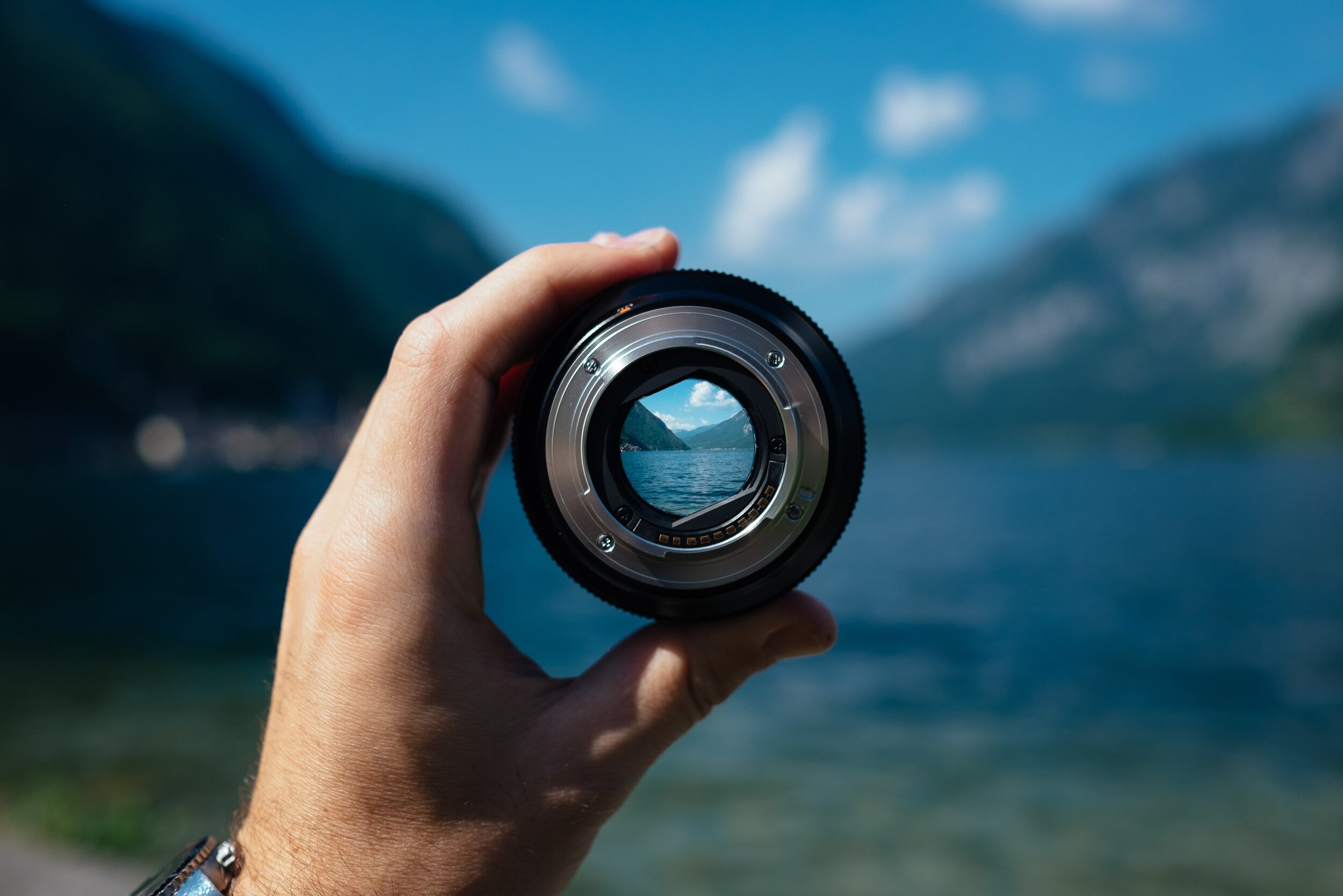 A hand holds a camera lens up to a lake and mountain landscape, framing the view within it