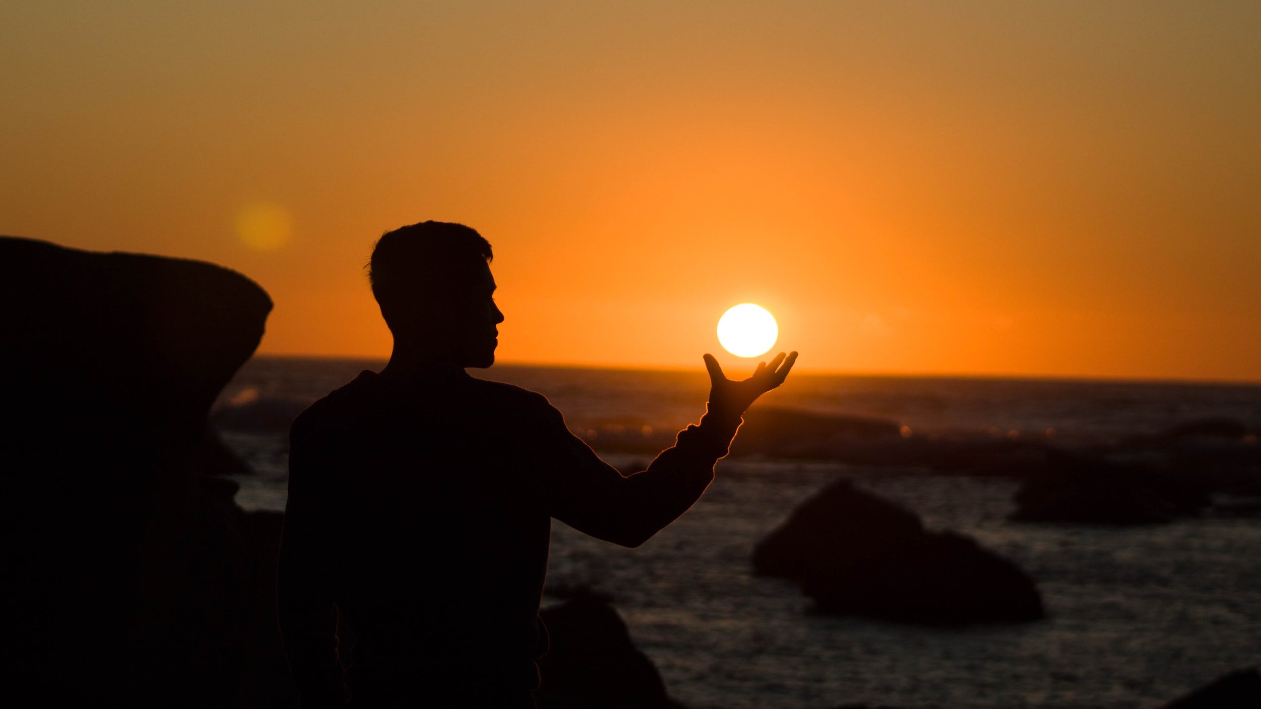 A silhouetted figure at sunset appears to hold the sun in an outstretched hand
