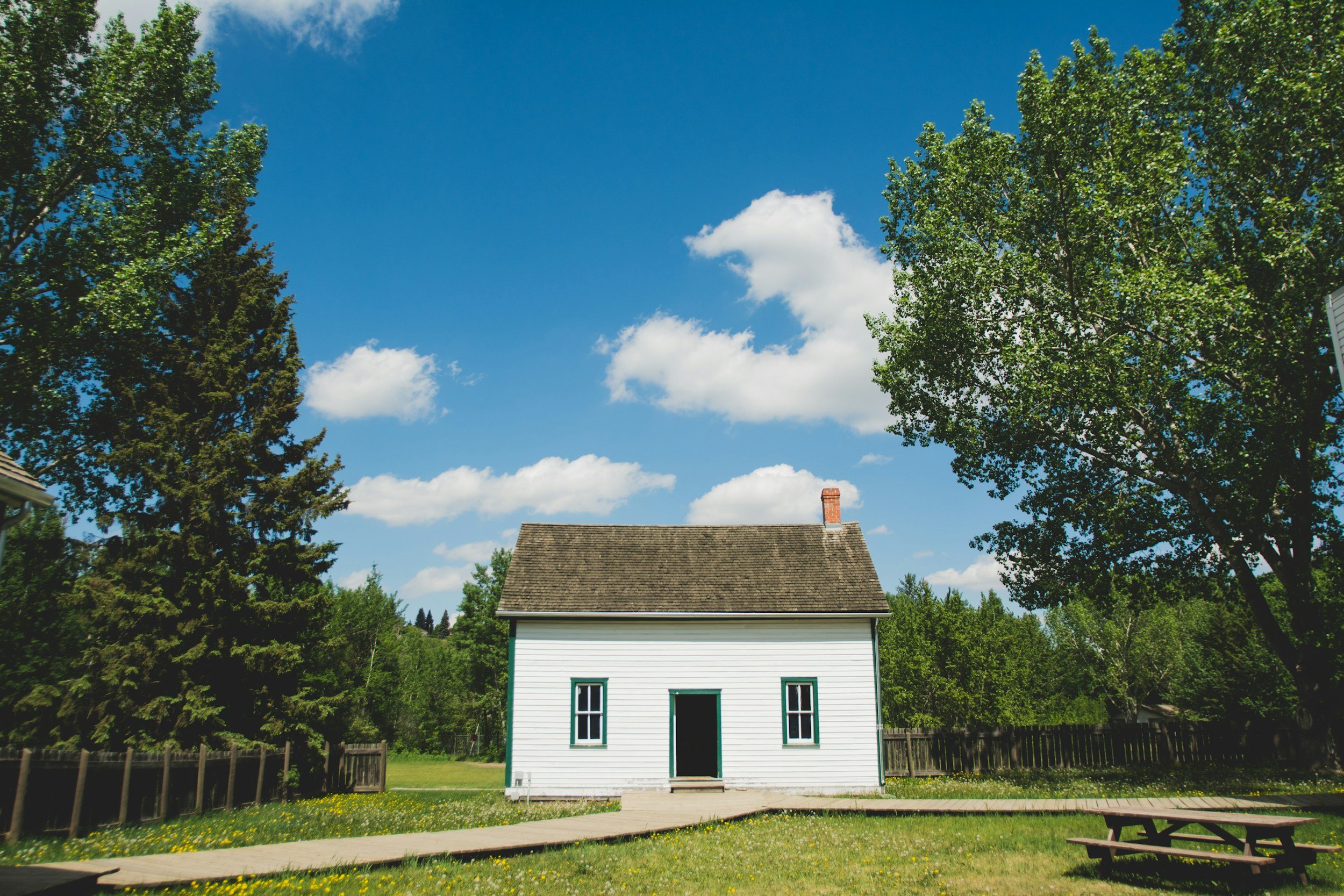 A small white house with green trim surrounded by trees under a blue sky